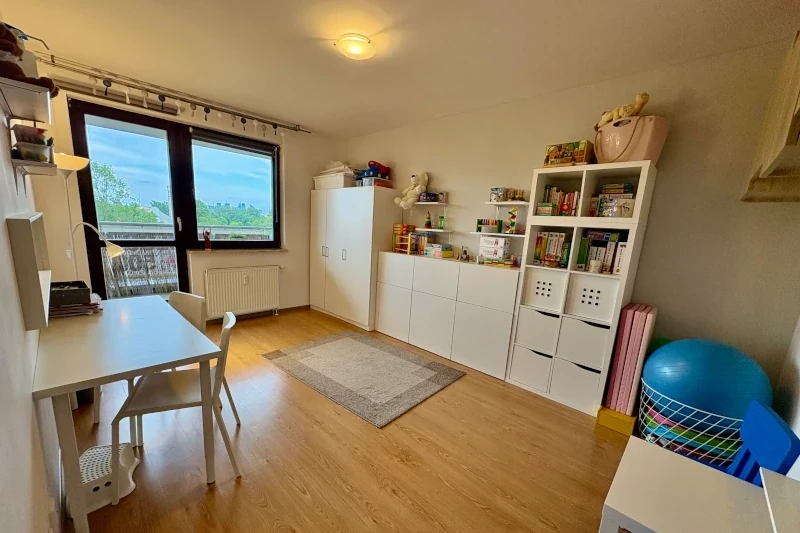 Bright children’s room with a large window, a white desk and chair, white storage cabinets with toys and books, a soft rug, and a blue exercise ball in the corner. The room has wooden floors and a tidy, organized appearance.