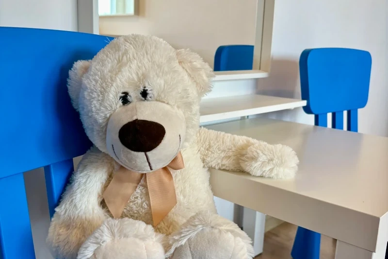 A fluffy white teddy bear with a beige ribbon around its neck is sitting on a blue chair next to a white children's table, with another blue chair and a mirror in the background.
