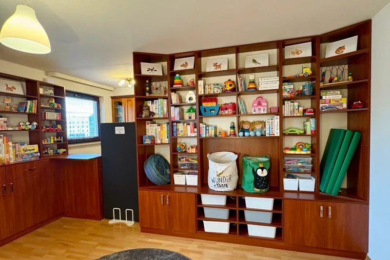 Spacious playroom with a large wooden shelving unit filled with children's books, board games, toys, and bins for storage. The room is well-organized and brightly lit, creating a welcoming environment for children.