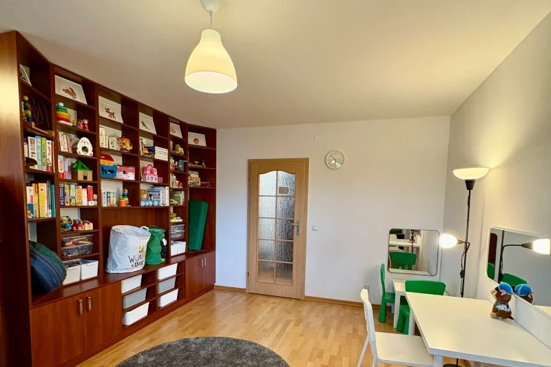 Children's playroom featuring a large wooden shelving unit filled with books, toys, and storage bins on the left wall. On the right side, there are small white tables with green chairs and mirrors, and the room is well-lit with overhead and floor lamps.