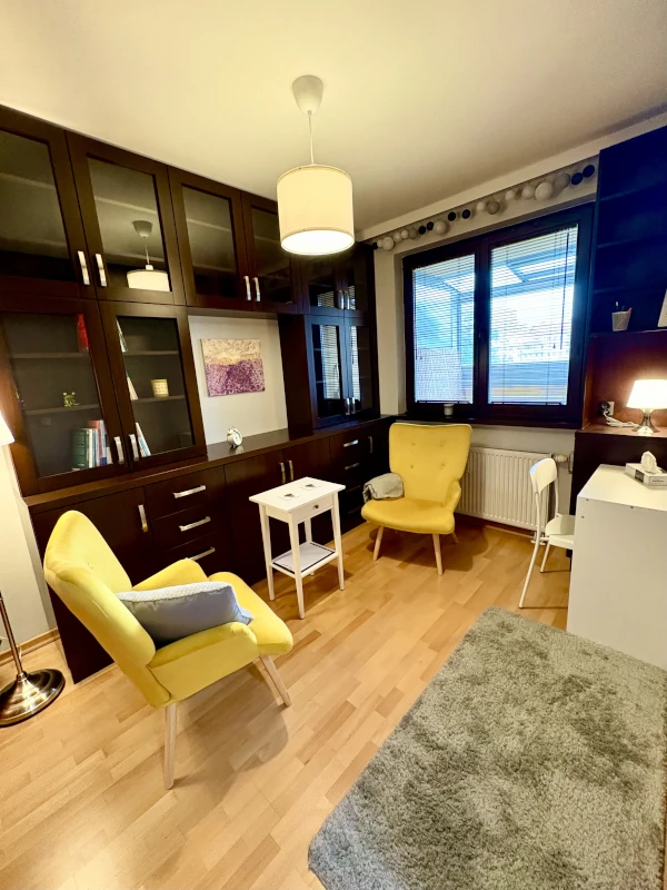 Cozy, well-lit office or reading room with two yellow armchairs, a small white table, and a large, dark wood bookshelf with glass cabinets. There is a gray rug on the wooden floor, a window with blinds, and a white desk with a lamp in the corner.