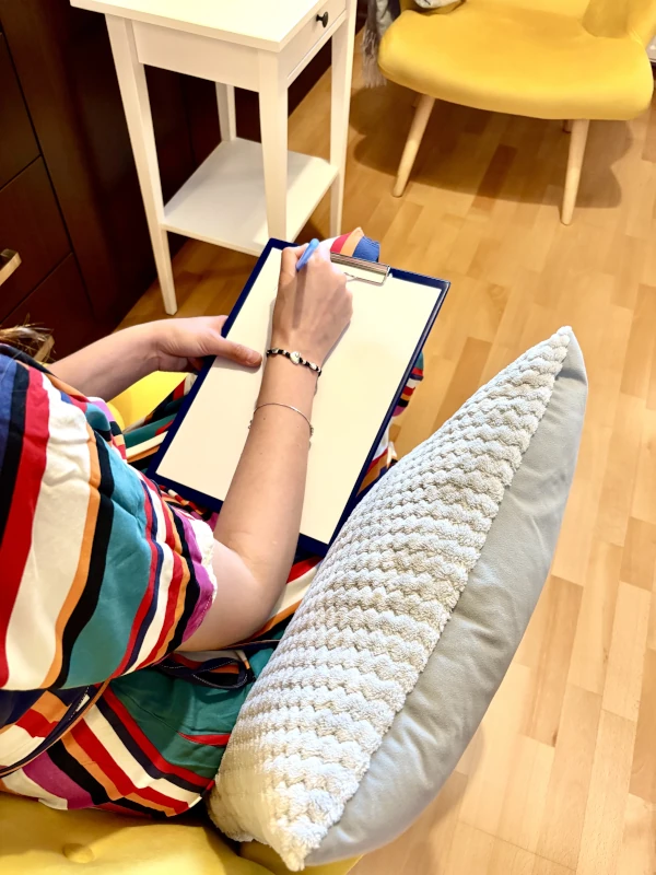Person wearing a colorful striped dress sits in a yellow chair with a textured cushion, writing on a blank sheet of paper attached to a blue clipboard. A small white side table and another yellow chair are visible in the background on a wooden floor.