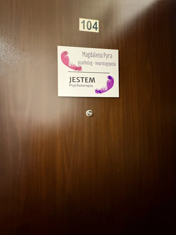 Brown wooden door with a room number 104 sign above a placard that reads 'Magdalena Pyra, psycholog - neurologopeda, JESTEM Psychoterapia' with two pink and purple feather graphics.