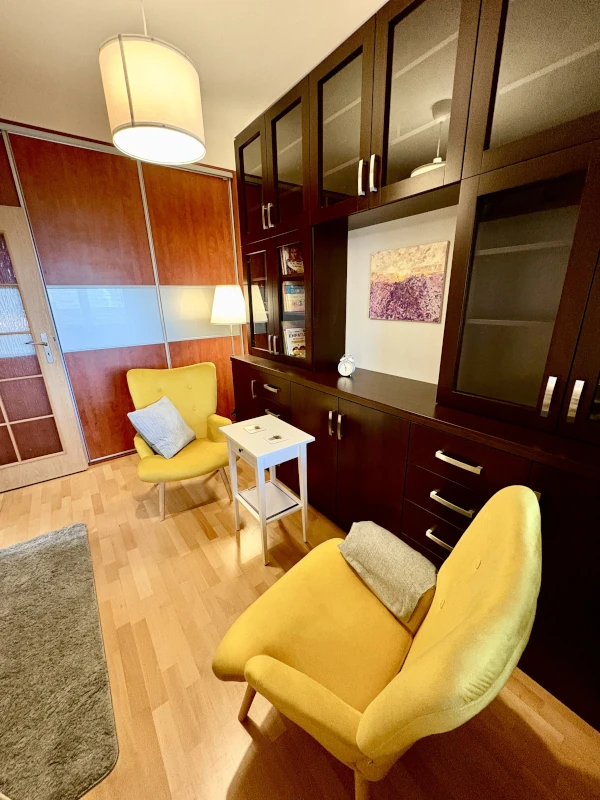 Comfortable office or therapy room with two yellow armchairs and gray pillows, a small white side table, and dark wood built-in shelving with glass cabinets. A floor lamp and a piece of purple abstract art decorate the space, which has wooden flooring and a soft gray rug.