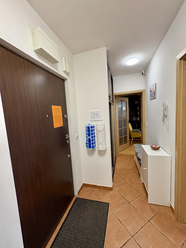 Entrance hallway with a dark brown door on the left, a tiled floor, and a white wall featuring dispensers for shoe covers. In the background, a doorway leads to a cozy room with two yellow chairs and dark wood shelving. A small white cabinet and a bowl are visible along the right wall.