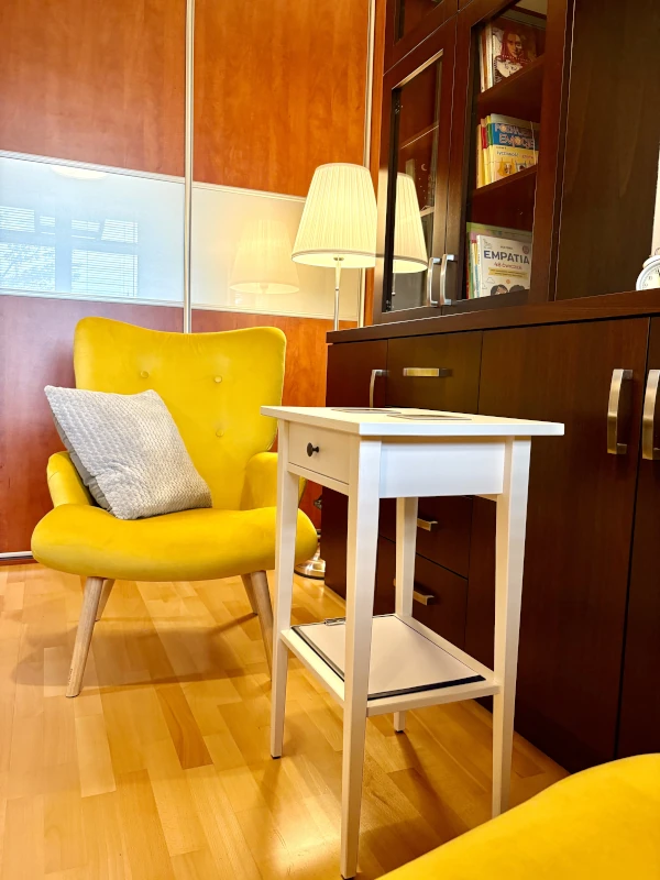 Cozy seating area with a bright yellow armchair accented by a gray pillow, a small white side table, and a dark wood cabinet with glass doors. A floor lamp and books are visible in the background, and the room features wooden flooring and sliding door panels.