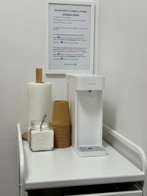 Hot water dispenser setup on a white cart, featuring a hot water dispenser, a stack of brown paper cups, a jar of sugar with a spoon, and a roll of paper towels. A framed instruction sheet in Polish is mounted on the wall behind the station.